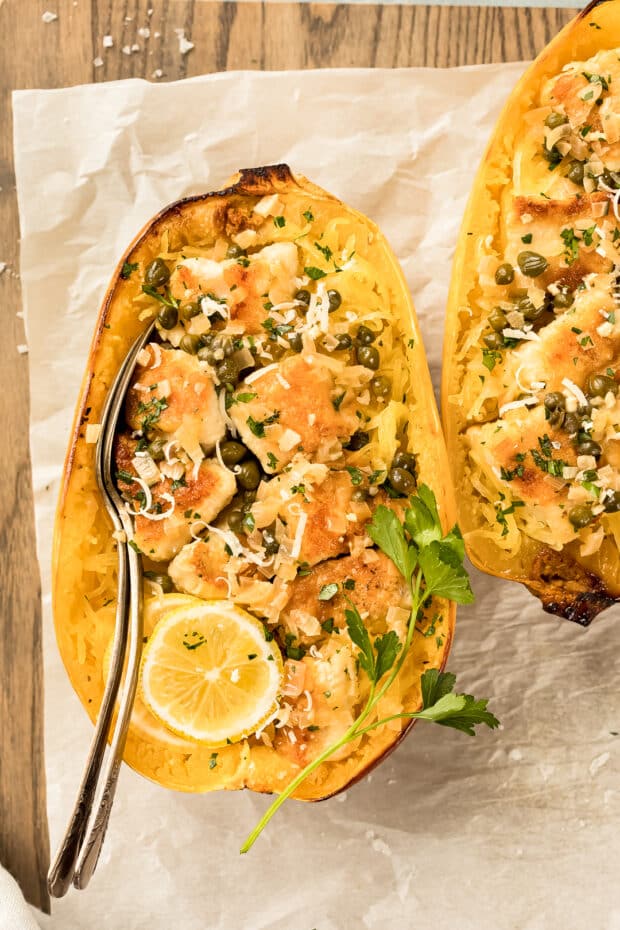 Overhead shot of Healthy Chicken Piccata Spaghetti Squash Boats garnished with a slice of lemon and sprig of fresh parsley with a fork and spoon inserted into the spaghetti squash boat with an additional chicken piccata boat next to it.