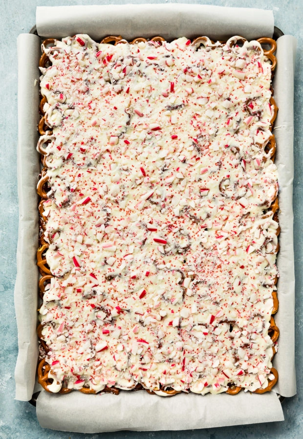 Overhead photo of a rimmed cookie sheet lined with parchment paper and filled with a layer of pretzels covered with toffee, white chocolate and crushed candy canes - photo of step 9 of the Bark recipe.
