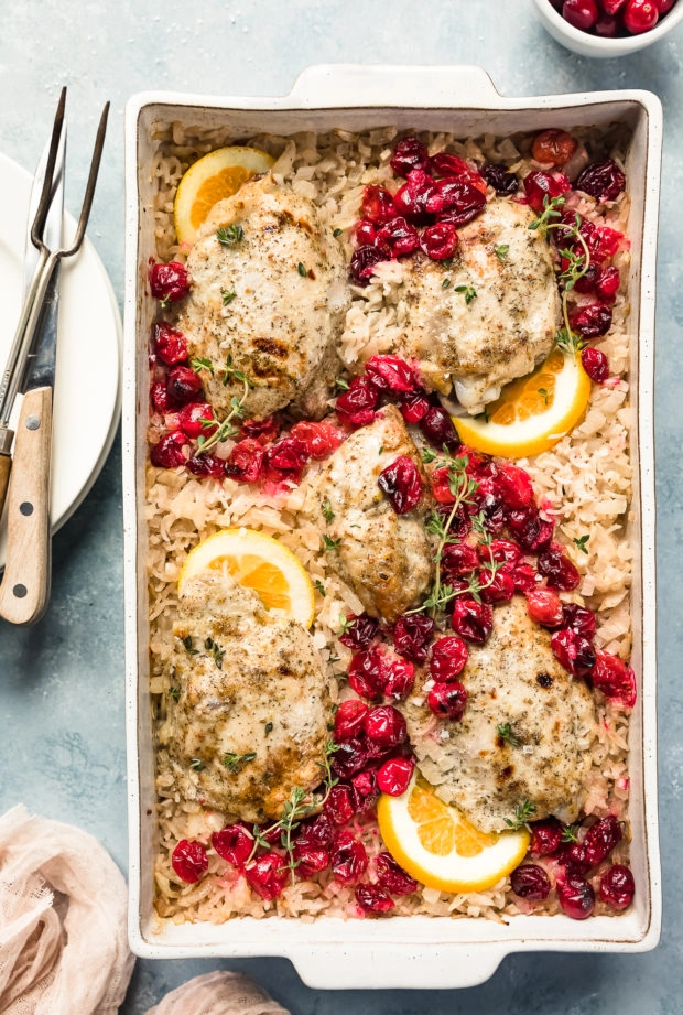 Overhead photo of Baked Orange Cranberry Chicken & Rice in a white casserole dish garnished with orange slices and fresh thyme with a ramekin of cranberries, neutral linen, stack of plates, knife and carving fork arranged around the dish.