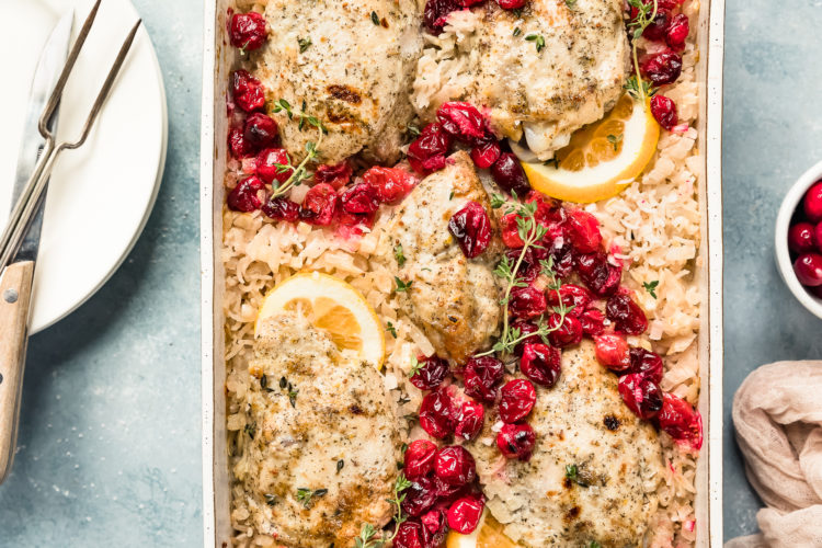 Overhead, landscape photo of Baked Orange Cranberry Chicken & Rice in a white casserole dish garnished with orange slices and fresh thyme with a ramekin of cranberries, neutral linen, stack of plates, knife and carving fork arranged around the dish.