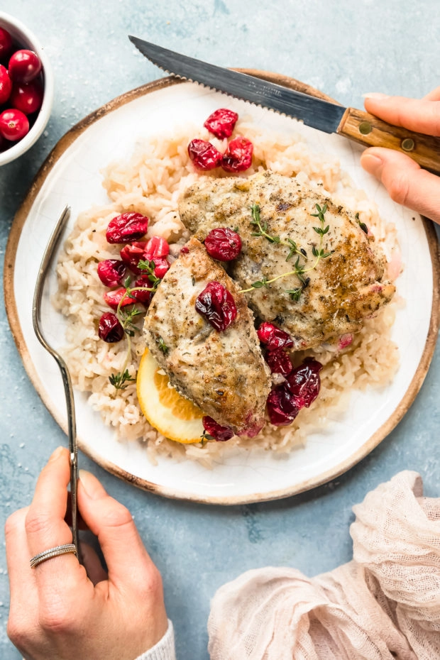 Overhead photo of Baked Orange Cranberry Chicken & Rice on a white plate with a pair of hands holding a knife and fork off to the sides of the plate.