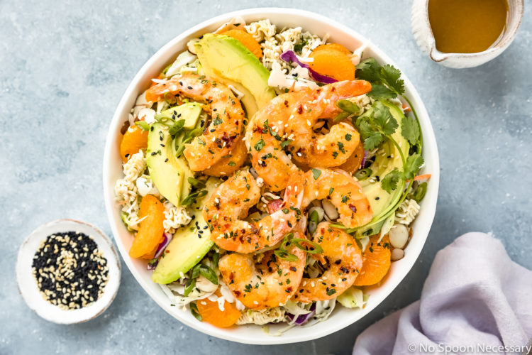 Overhead photo of Healthy Orange Shrimp Ramen Noodle Salad with coleslaw and sliced avocado in a white bowl with a ramekin of sesame seeds, a purple linen and a small jar of Asian dressing surrounding the bowl.