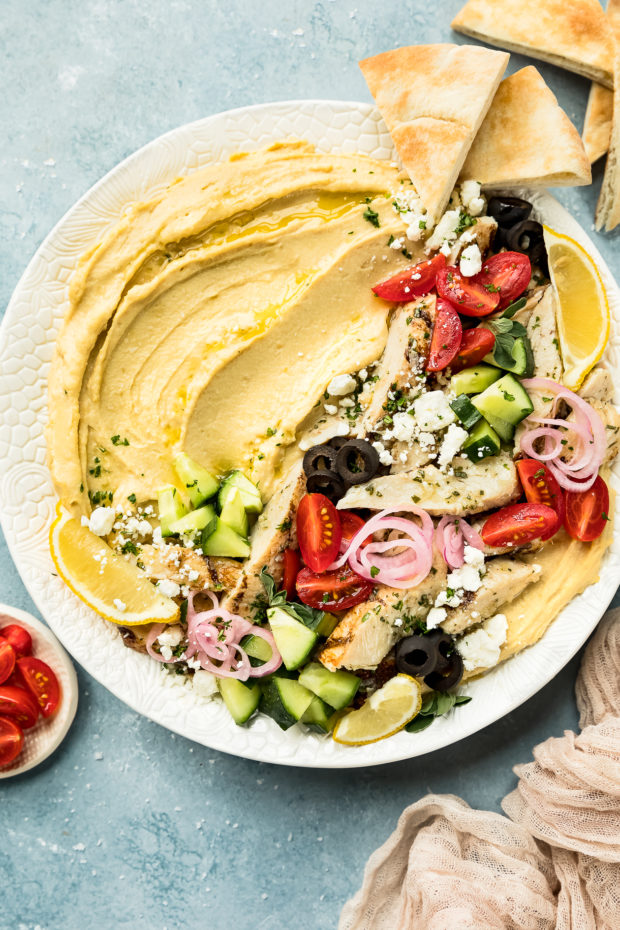 Overhead photo of a Mediterranean Hummus Bowl topped with chicken and vegetables on a large white platter with pita triangles and a ramekin of halved cherry tomatoes next to the platter.