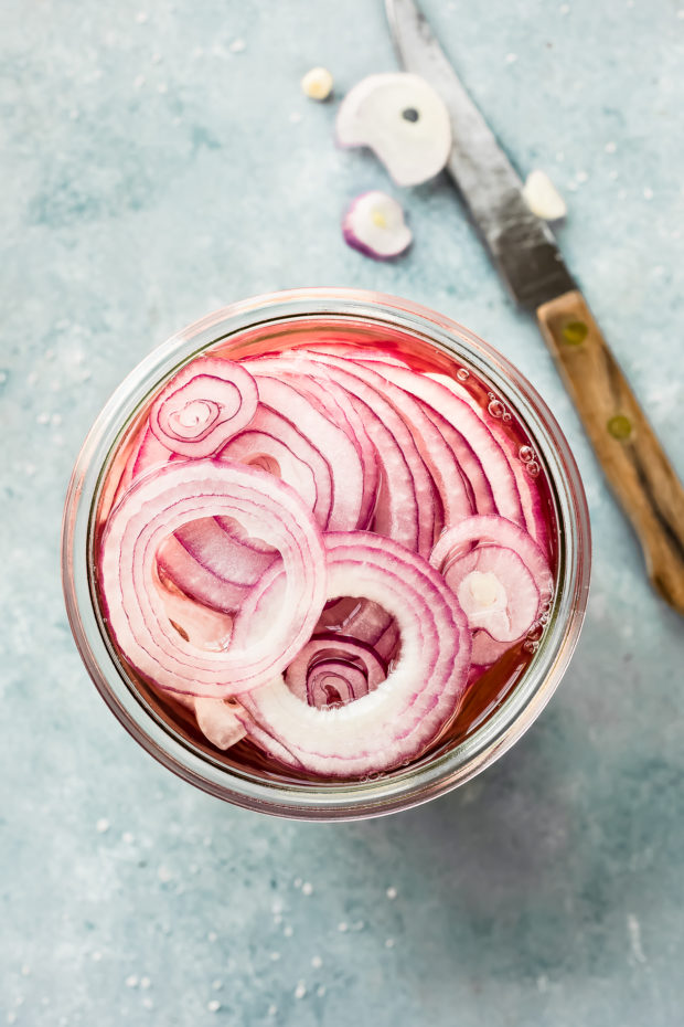Overhead photo of a large jar of thinly sliced red onions in a pickling brine with a knife next to the jar - photo of onion prep for Hummus Bowls.