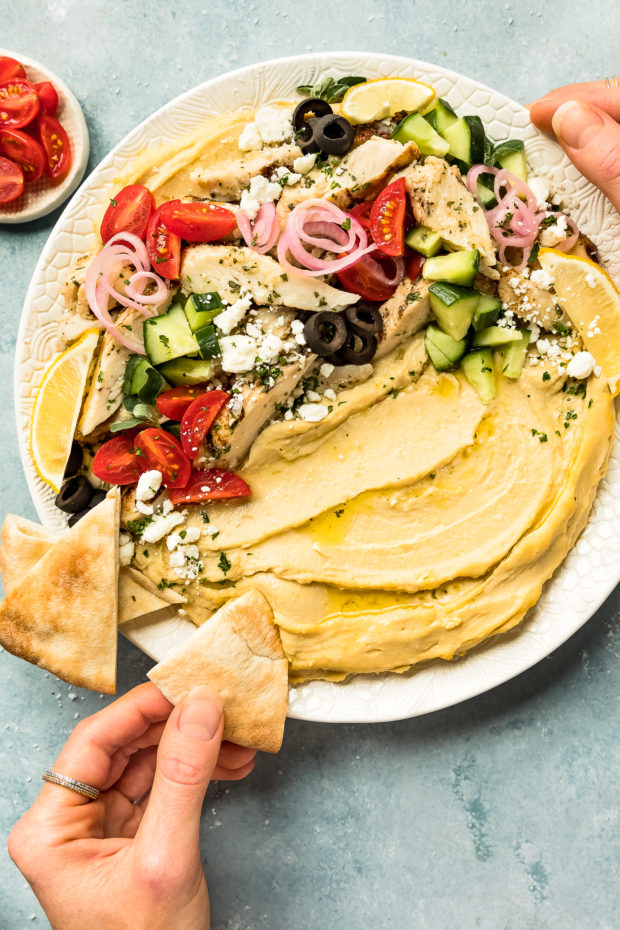 Overhead photo of a Chicken Hummus Bowl on a large white platter with a hand dipping a pita triangle into the hummus and a ramekin of halved cherry tomatoes next to the platter.