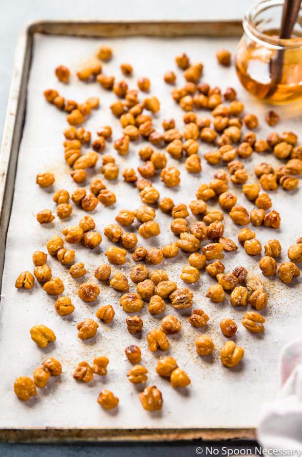 Angled photo of Crispy Honey Roasted Chickpeas dusted with ground ginger on a small parchment paper lined sheet pan with a glass jar of honey on the pan and a pale purple linen next to the pan.