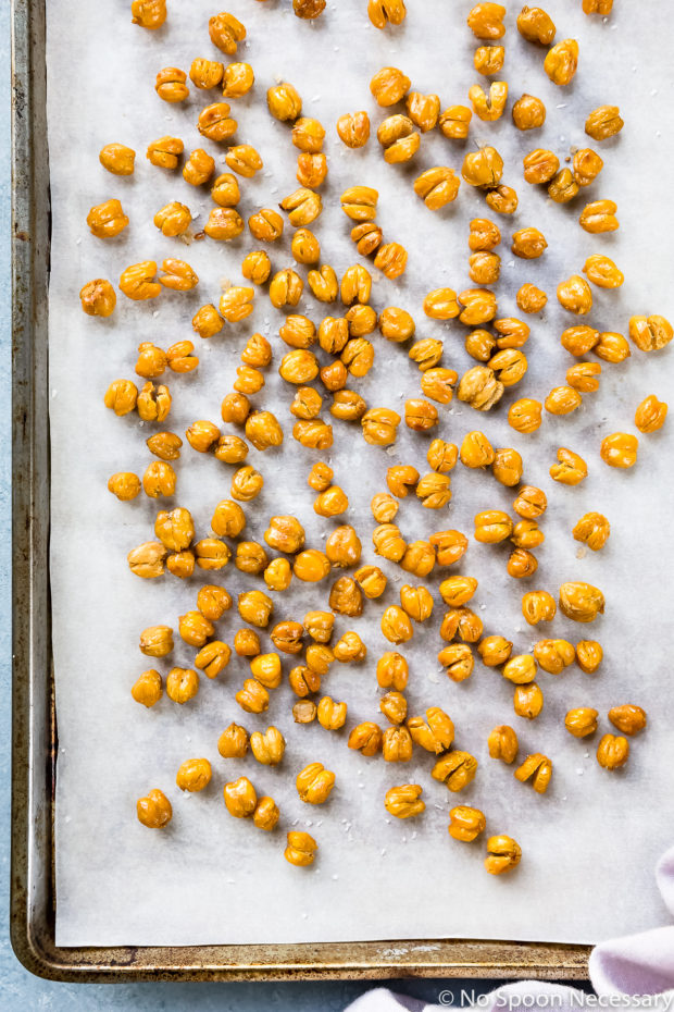 Overhead photo of roasted chickpeas on a parchment paper lined sheet pan before being tossed with honey - photo of step 4 of the Crispy Honey Roasted Chickpeas recipe.