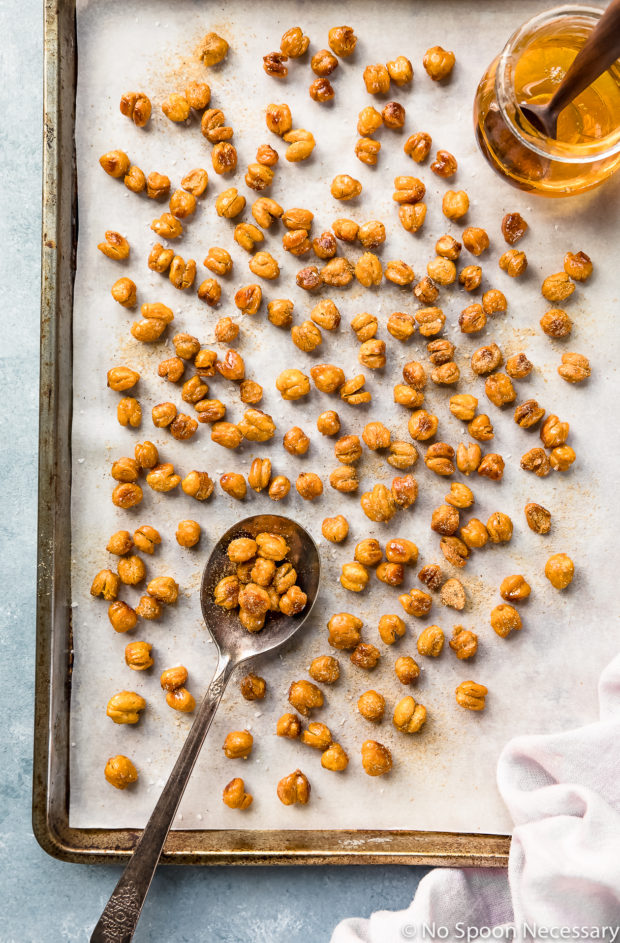 Overhead photo of Crispy Honey Roasted Chickpeas on a small parchment paper lined sheet pan with a glass jar of honey on the pan and a pale purple linen next to the pan.