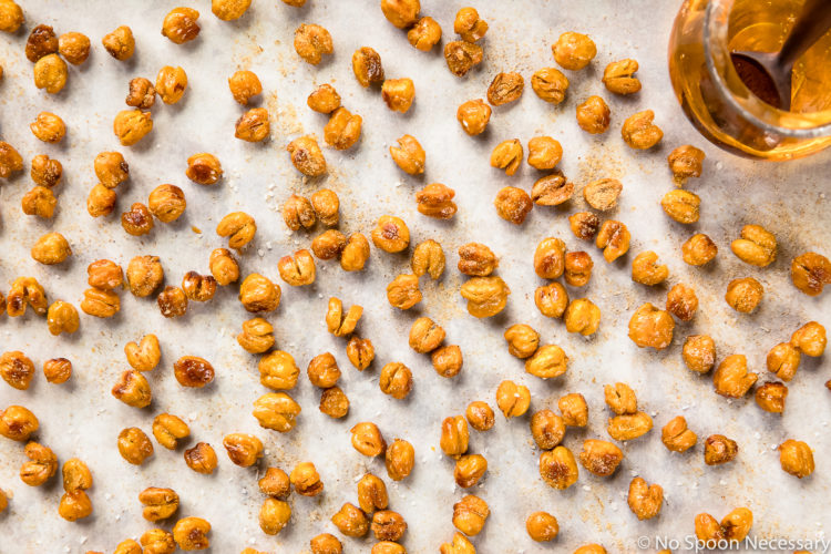 Overhead, up-close photo of Crispy Honey Roasted Chickpeas dusted with ground ginger and sprinkled with flaky sea salt on a piece of parchment paper with a glass jar of honey in the corner of the shot.