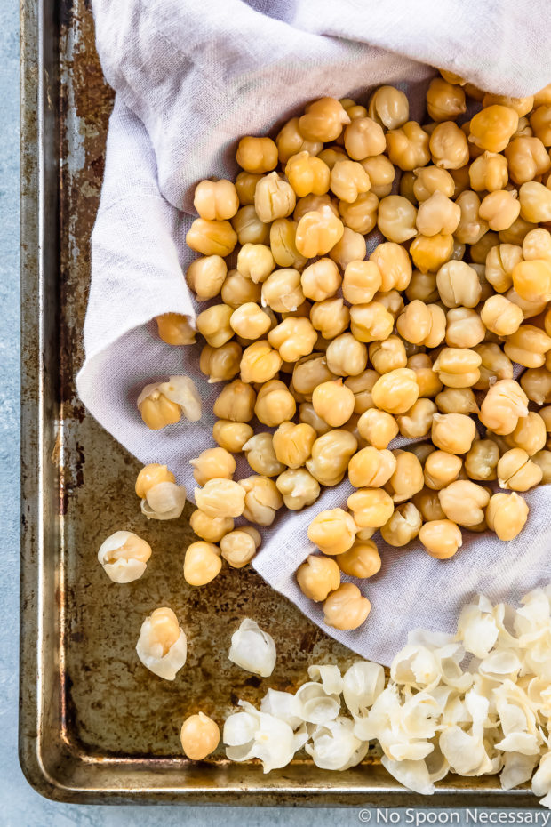 Overhead photo of cooked chickpeas on a pale purple linen lined sheet pan with half of the skins removed from the chickpeas - photo of how to remove chickpea skins to make Crispy Honey Roasted Chickpeas recipe.