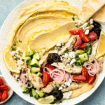 Overhead photo of a Hummus Bowl topped with chicken and fresh vegetables on a large white platter with pita triangles and a ramekin of halved cherry tomatoes and a jar of Greek vinaigrette next to the platter.
