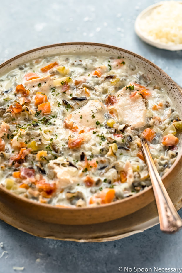 Angled photo of Slow Cooker Creamy Chicken and Wild Rice Soup in a neutral colored bowl on a tan plate with a spoon inserted into the soup and a ramekin of shredded parmesan blurred in the background.