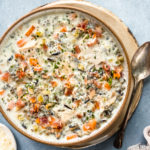 Overhead, landscape photo of Slow Cooker Creamy Chicken and Wild Rice Soup in a neutral colored bowl on a tan plate with a spoon resting next to the bowl and a ramekin of shredded parmesan, white wine glass and pale tan linen arranged around the bowl.