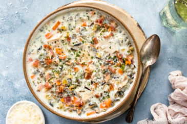 Overhead, landscape photo of Slow Cooker Creamy Chicken and Wild Rice Soup in a neutral colored bowl on a tan plate with a spoon resting next to the bowl and a ramekin of shredded parmesan, white wine glass and pale tan linen arranged around the bowl.
