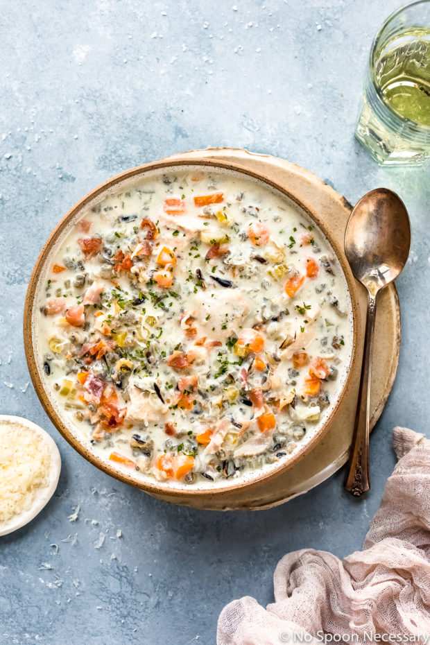 Overhead photo of Slow Cooker Creamy Chicken and Wild Rice Soup in a neutral colored bowl on a tan plate with a spoon resting next to the bowl and a ramekin of shredded parmesan, white wine glass and pale tan linen arranged around the bowl.