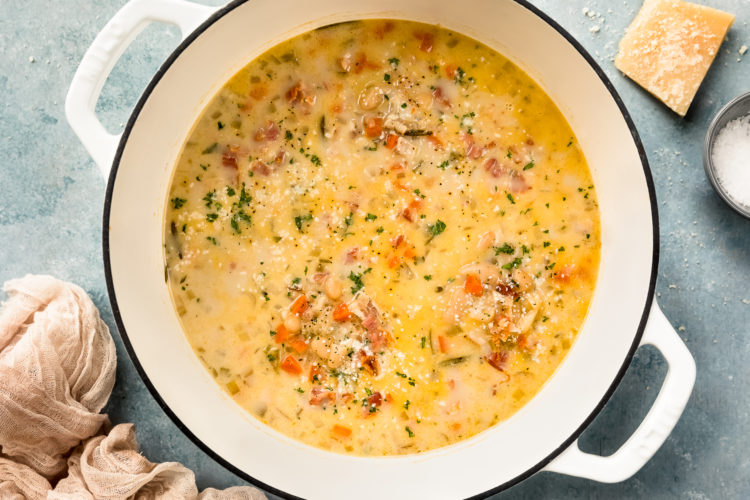 Overhead photo of a large white pot filled with Tuscan White Bean Soup with bacon with a chunk of parmesan and ramekin of salt next to the pot.