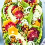 Overhead photo of Winter Citrus Salad with avocado, cucumbers and mint on a large white platter with a pale purple linen and ramekins of honey citrus vinaigrette and crispy chickpeas surrounding the platter.