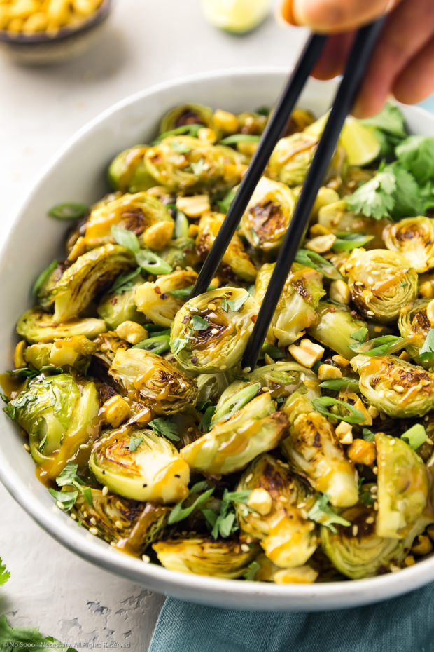 Angled photo of Asian Brussel Sprouts drizzled with peanut sauce in a white serving bowl with a hand holding a pair of black chop stick picking up an individual sprout