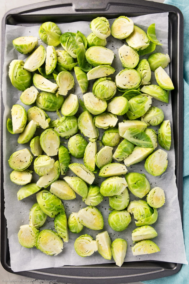 Overhead photo of trimmed and halved raw brussels sprouts seasoned with salt and pepper on a parchment paper lined sheet pan - photo of step 2 of the recipe.