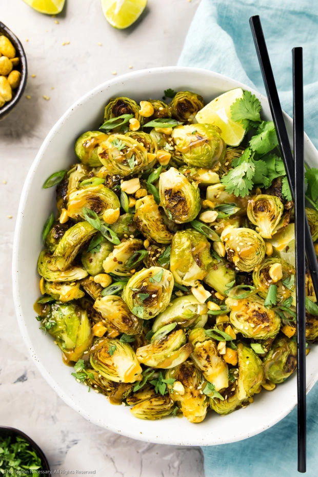 Overhead photo Crispy Asian Brussel Sprouts drizzled with peanut sauce in a white serving bowl with a pale blue linen, ramekin of peanuts, ramekin of sliced scallions and lime wedges arranged around the bowl.