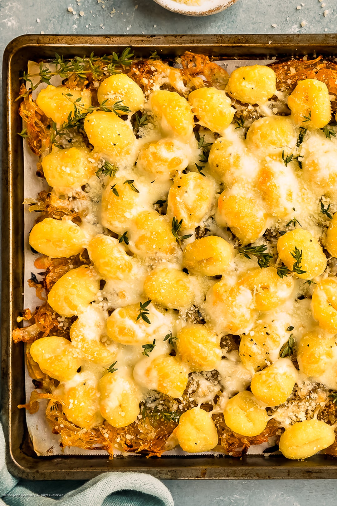 Overhead photo of cheesy baked gnocchi with onions and fresh herbs on a baking sheet.