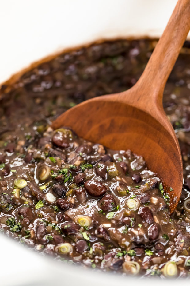 Angled, up-close photo of cooked Cuban Black Beans in a large white dutch with a wooden serving spoon inserted into the pot.