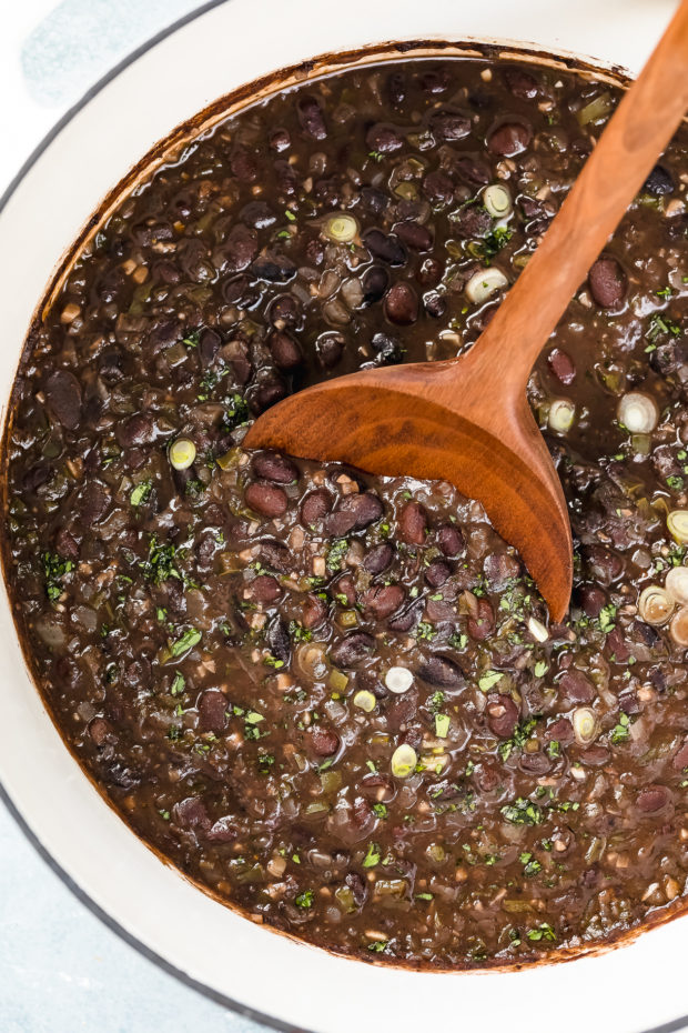 Overhead, up-close photo of cooked Cuban Black Beans in a large white dutch oven garnished with fresh cilantro and sliced scallions with a wooden serving spoon inserted into the pot.