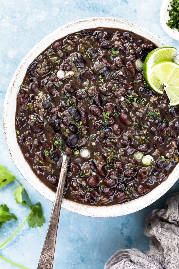Overhead photo of Cuban Black Beans in a neutral colored serving bowl with a spoon inserted into the beans and lime wedges garnishing the top.