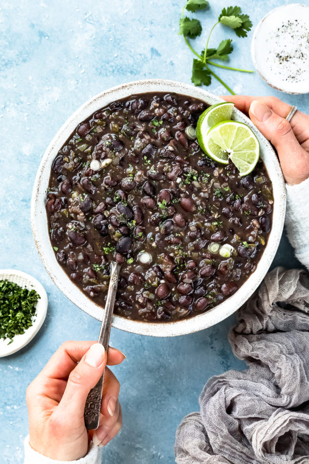 Overhead photo of Cuban Black Beans in a neutral colored serving bowl with a hand holding a spoon inserted into the beans and ramekins of chopped cilantro and salt arranged around the bowl.