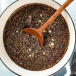 Overhead photo of cooked Cuban Black Beans in a large white dutch oven garnished with fresh cilantro and sliced scallions; with a ramekin of chopped cilantro and salt arranged around the pot.