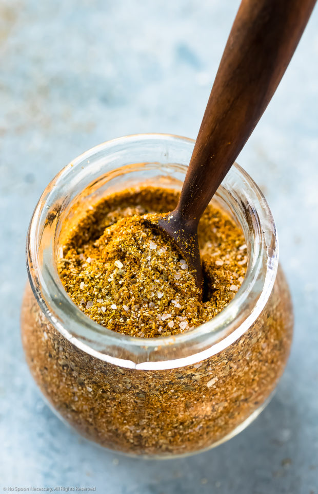 Angled, up-close photo of Homemade Sazon Seasoning Mix in a small glass jar with a small wooden spoon inserted into the jar.