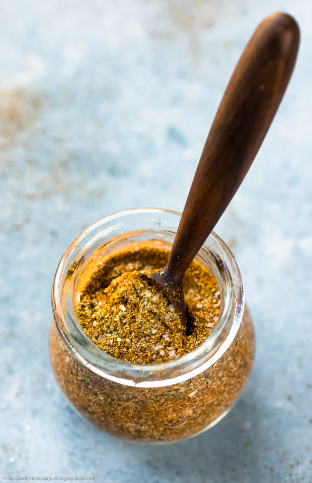 Angled photo of Homemade Sazon Seasoning Mix in a small glass jar with a small wooden spoon inserted into the jar.