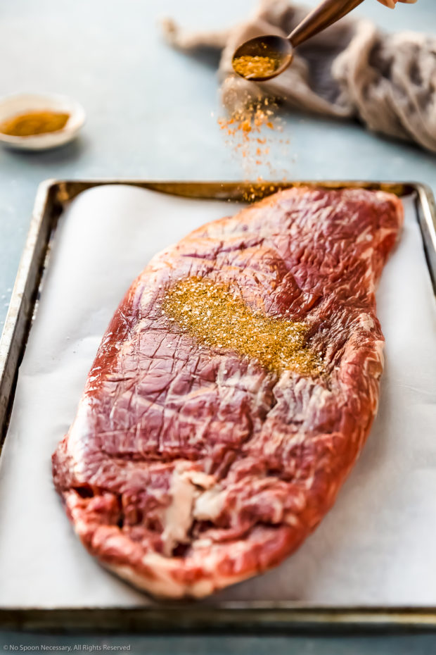 Angled photo of Sazon seasoning being sprinkled over flank steak on a parchment paper lined sheet pan - photo of step 1 of the Slow Cooker Ropa Vieja recipe.