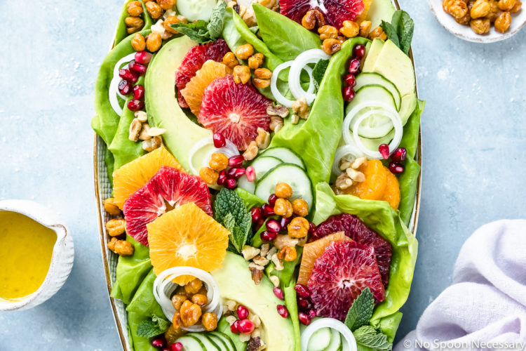 Overhead photo of Winter Citrus Salad with avocado, cucumbers and mint on a large white platter with a pale purple linen and ramekins of honey citrus vinaigrette and crispy chickpeas surrounding the platter.