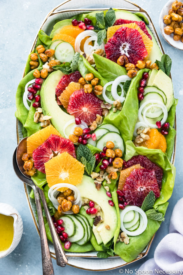 Overhead photo of Winter Citrus Salad with avocado, cucumbers and mint on a large white platter with serving spoons tucked into the salad and a pale purple linen and ramekins of honey citrus vinaigrette and crispy chickpeas surrounding the platter. 