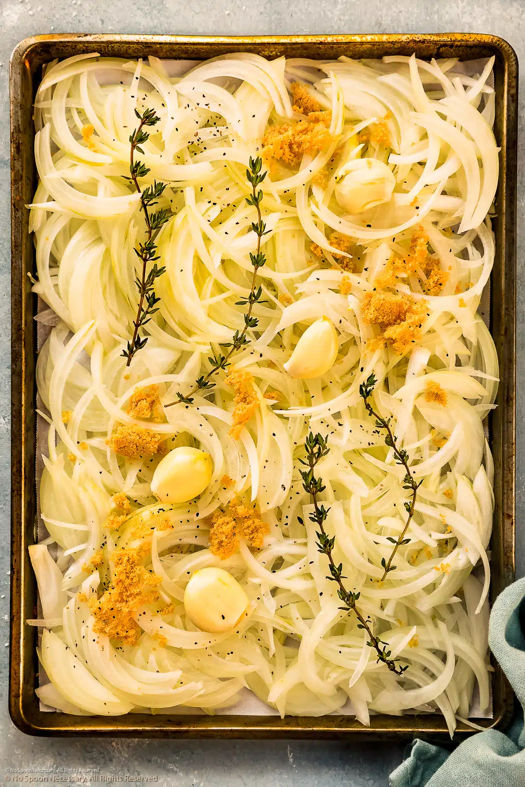 Overhead photo of thinly sliced yellow onions with three cloves of garlic, brown sugar, and fresh thyme sprigs on a sheet pan - before baking.