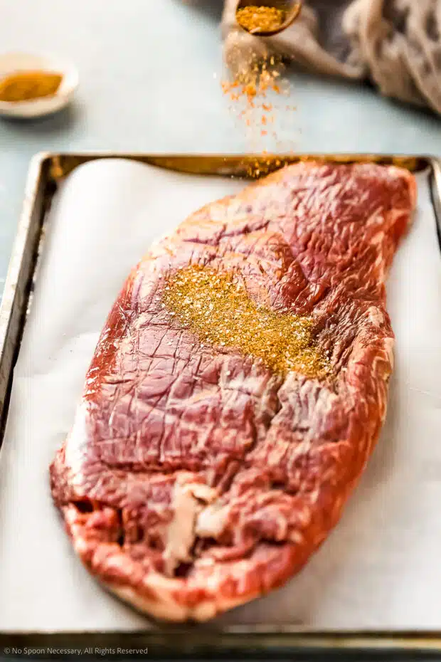 Angled photo of Sazon seasoning being sprinkled over flank steak on a parchment paper lined sheet pan.