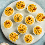Overhead, landscape photo of Bacon Deviled Eggs garnished with a slice of bacon on a white plate with a ramekin of snipped chives and a pale tan napkin next to the plate.