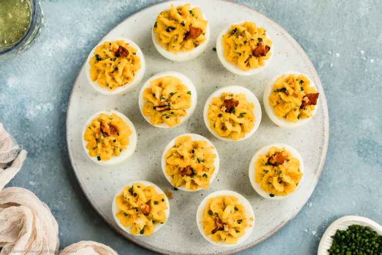 Overhead, landscape photo of Bacon Deviled Eggs garnished with a slice of bacon on a white plate with a ramekin of snipped chives and a pale tan napkin next to the plate.