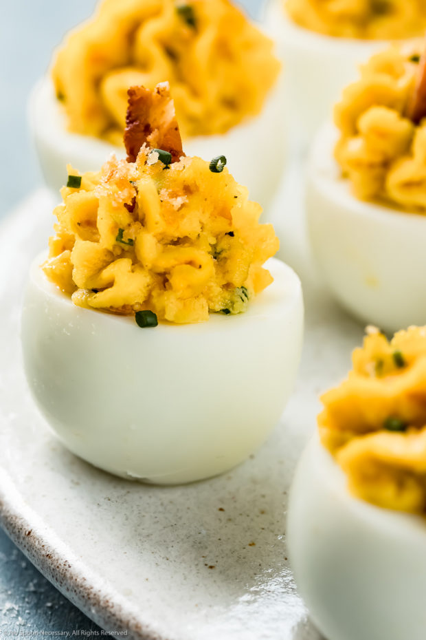 Angled photo of Cheddar and Bacon Deviled Eggs garnished with a slice of bacon and snipped chives on a white plate.