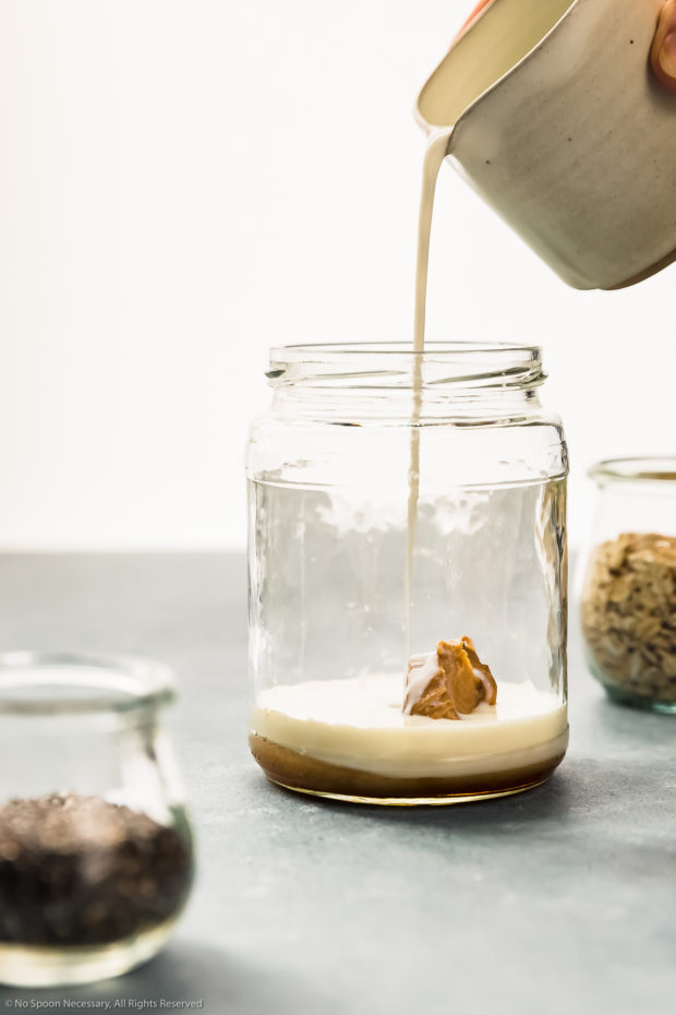 Straight on photo of milk being poured from a spouted container into a glass jar containing peanut butter and maple syrup - photo of the first part of step 1 of the recipe.