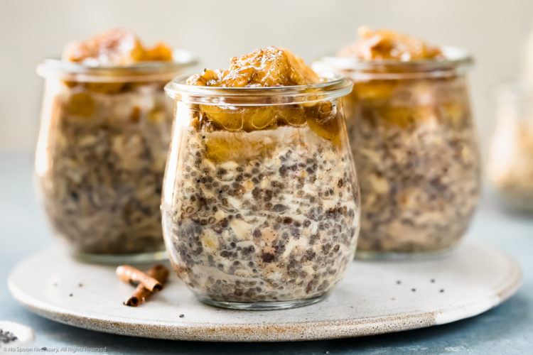 Straight on photo of three individual jars of Banana Overnight Oats on a white plate with spoons next to the jars and a glass jar of rolled oats blurred in the background.