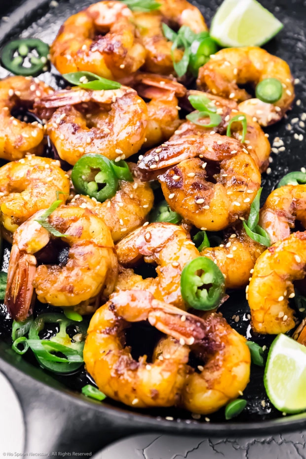 Angled, overhead photo of Garlic Honey Shrimp garnished with lime wedges, jalapeno slices, and sliced scallions in a large cast iron skillet