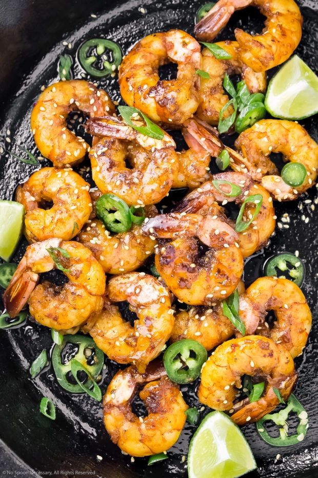 Overhead shot of a cast iron skillet filled with Sticky Honey Garlic Glazed Shrimp, lime wedges, jalapeno slices, and sliced scallions.