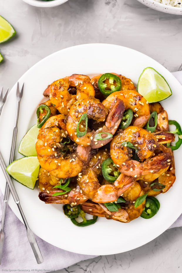 Overhead photo sweet and spicy Honey Shrimp garnished with lime wedges, jalapeno slices and sliced scallions on a white plate with a purple linen under the plate and a bowl of rice next to the plate.