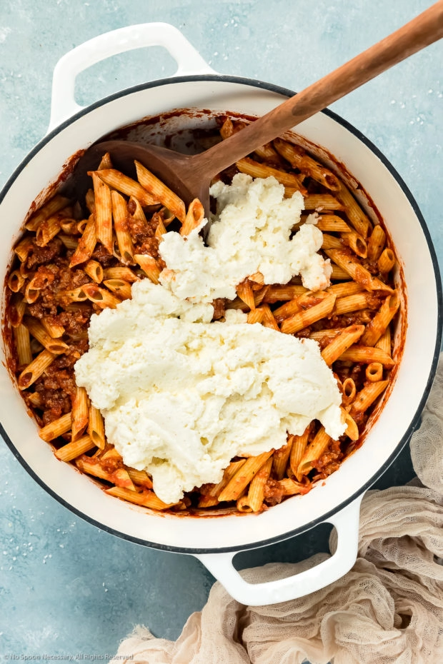 Overhead photo of soaked ziti noodles tossed with sausage tomato sauce and topped with ricotta in a large white pot with a wooden spoon inserted into the pot - photo of step 9 of the recipe.