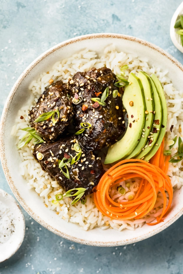 Overhead shot of Slow Cooker Korean Short Ribs on a bed of rice with sliced avocado and carrot ribbons in a white bowl with ramekins of salt and sliced scallions arranged around the bowl.