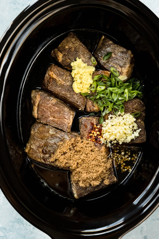Overhead photo of a black slow cooker insert filled with seared short ribs, brown sugar, garlic, ginger, scallions, red pepper flakes and Korean sauce - photo of step 3 of the recipe.
