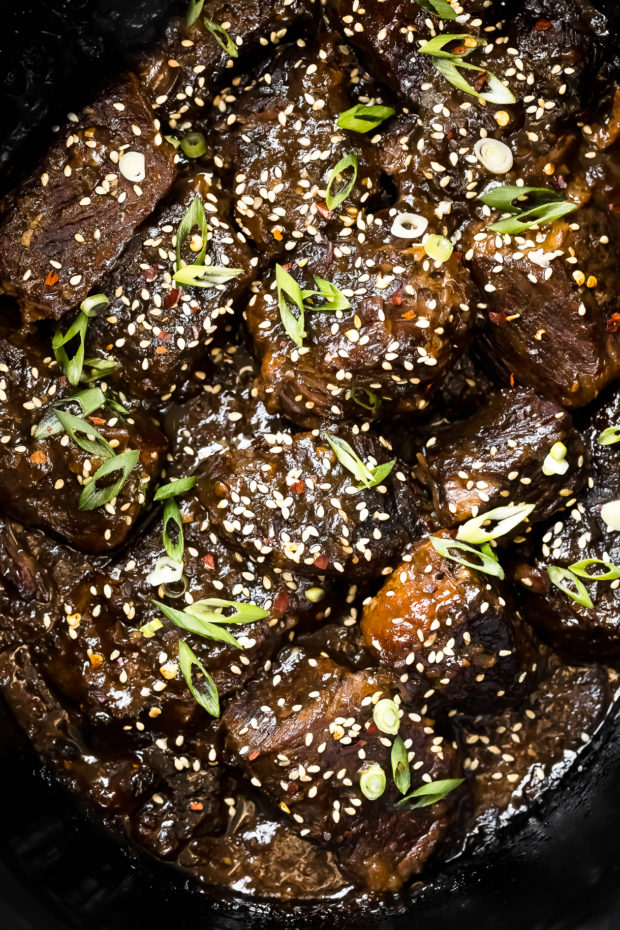 Overhead, photo of a black slow cooker filled with cooked boneless Asian Beef Short Ribs garnished with sliced scallions and sesame seeds.