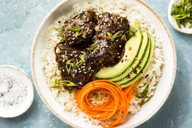 Overhead, landscape shot of Slow Cooker Korean Short Ribs on a bed of rice with sliced avocado and carrot ribbons in a white bowl with ramekins of salt and sliced scallions arranged around the bowl.